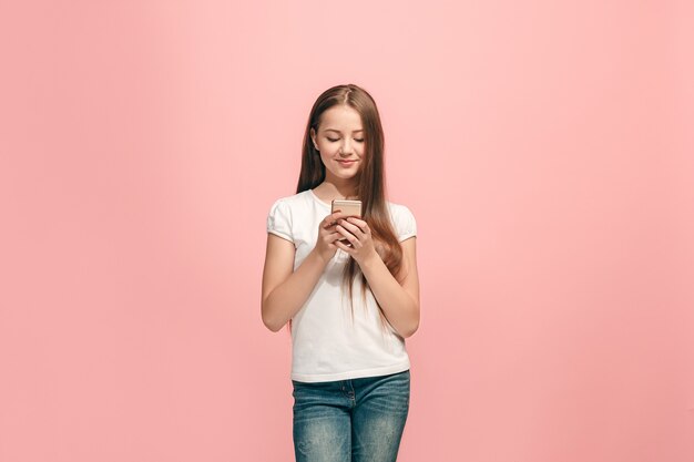 Felice ragazza adolescente in piedi, sorridente con il telefono cellulare sul muro rosa alla moda dello studio