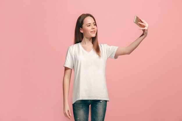 Felice ragazza adolescente in piedi, sorridente in studio rosa, facendo selfie foto dal telefono cellulare.