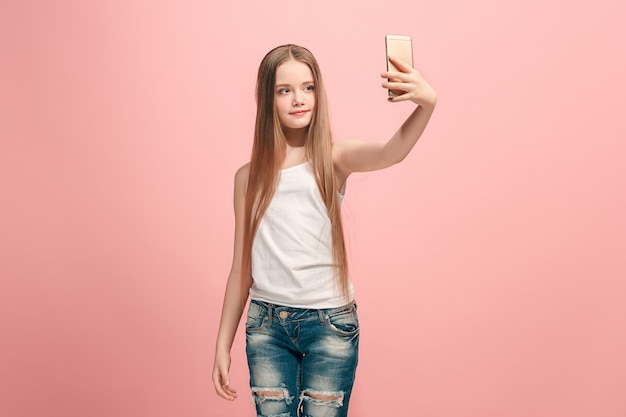 Felice ragazza adolescente in piedi, sorridente sul muro rosa, facendo selfie foto dal telefono cellulare. Emozioni umane, concetto di espressione facciale. Vista frontale.