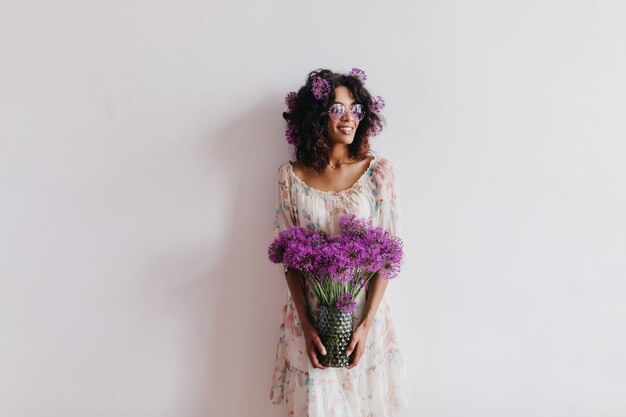 Felice ragazza africana con vaso di fiori e sorridente. Ritratto dell'interno della donna castana affascinante in vestito che posa con gli allium.