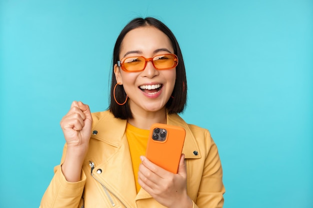 Felice ragazza asiatica alla moda utilizzando smartphone e ridendo sorridendo alla telecamera in piedi su sfondo blu