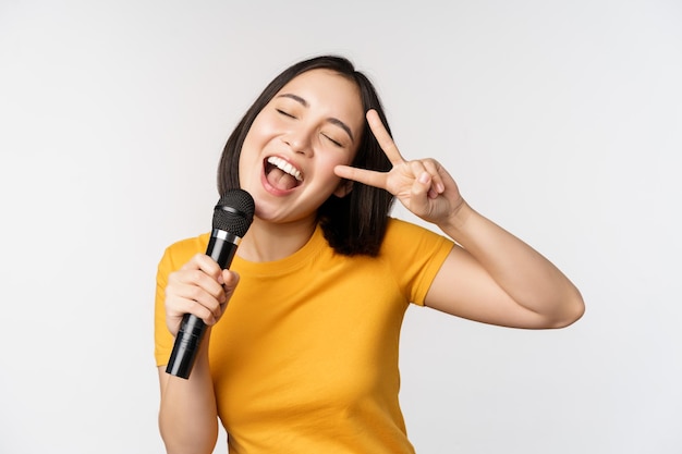 Felice ragazza asiatica ballare e cantare karaoke tenendo il microfono in mano divertendosi in piedi su sfondo bianco