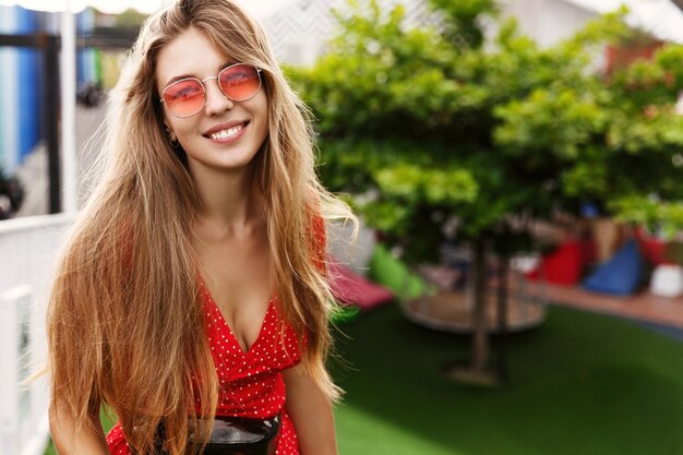 Felice ragazza bionda godersi le vacanze tropicali, in piedi nel parco verde e soleggiato e sorridente, indossando occhiali da sole rossi