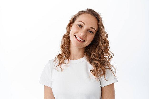 Felice ragazza candida con capelli biondi naturali make up sorriso e sguardo positivo alla testa di inclinazione della fotocamera carino si erge su sfondo bianco Spazio di copia