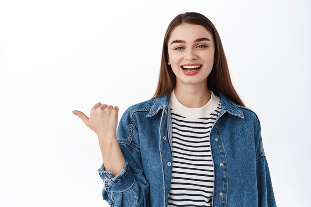 Felice ragazza candida in giacca estiva in denim, indicando da parte, ridendo e sorridendo compiaciuta, mostrando banner promozionale, testo su spazio copia o logo, in piedi sul muro bianco