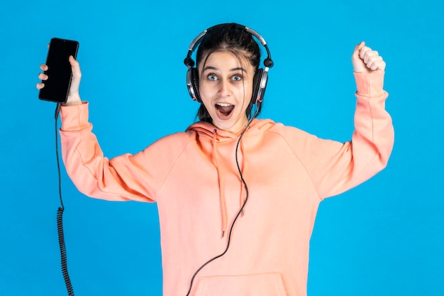 Felice ragazza che indossa le cuffie e tiene il suo telefono su sfondo blu Foto di alta qualità