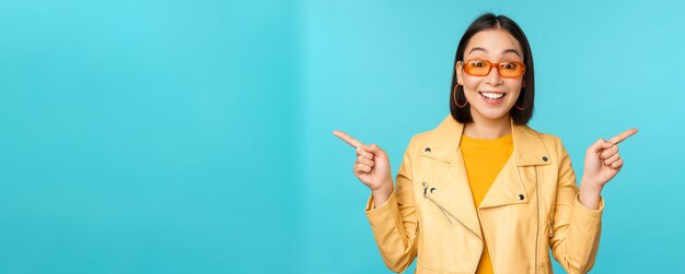 Felice ragazza cinese alla moda in occhiali da sole punta le dita lateralmente invita a controllare vedere lo sconto del negozio mostra stand sinistro e destro su sfondo blu