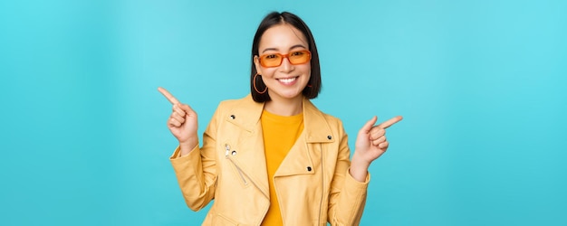 Felice ragazza cinese alla moda in occhiali da sole punta le dita lateralmente invita a controllare vedere lo sconto del negozio mostra stand sinistro e destro su sfondo blu