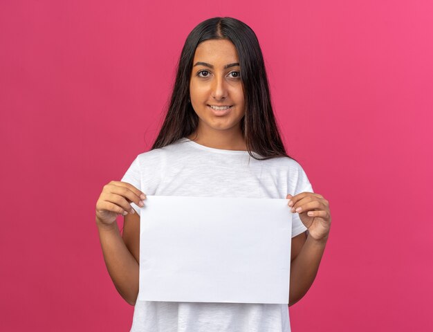 Felice ragazza con una maglietta bianca che tiene un foglio di carta bianco bianco che guarda la telecamera sorridendo allegramente