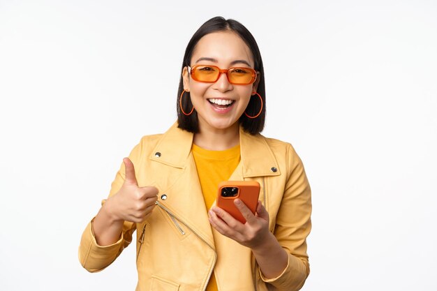 Felice ragazza coreana alla moda che indossa occhiali da sole che mostra il pollice in su tenendo lo smartphone utilizzando l'app del telefono cellulare consigliando l'applicazione negozio online sfondo bianco