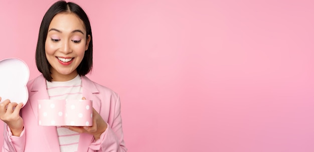 Felice ragazza coreana carina in tuta apre una scatola a forma di cuore con un regalo romantico in vacanza di un giorno bianco in piedi in tuta su sfondo rosa