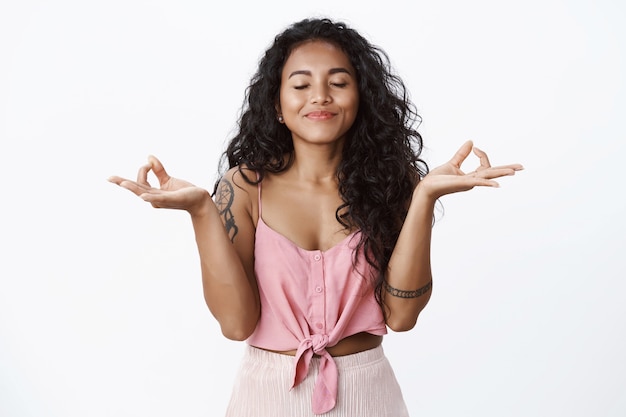 Felice ragazza dai capelli ricci con tatuaggi che meditano, alza le mani nel gesto zen, pratica lo yoga, chiudi gli occhi e sorride felice, inspira aria fresca, calmati sollevato, muro bianco