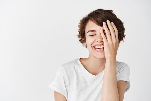 Felice ragazza europea con i capelli corti coprono metà del viso e sorridente che ride spensierata in piedi in maglietta su sfondo bianco