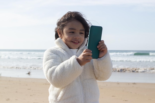 Felice ragazza giapponese con il cellulare sulla spiaggia. Bambino dai capelli scuri che tiene dispositivo e guarda lo schermo. Infanzia, concetto di tecnologia