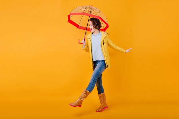 Felice ragazza riccia in jeans divertente ballare tenendo ombrellone alla moda. Ritratto in studio di giovane donna ispirata in scarpe di gomma che scherza sul muro giallo brillante e sorridente.