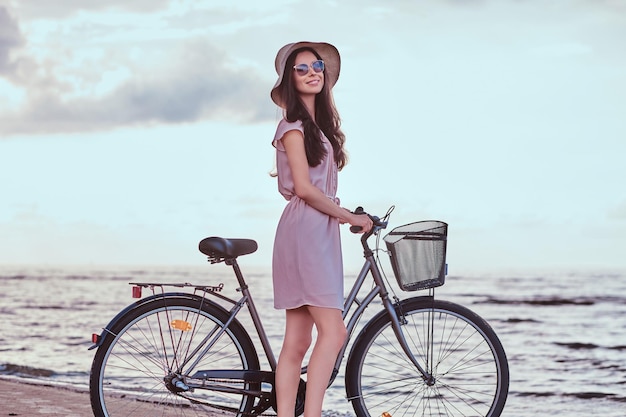 Felice ragazza sensuale in occhiali da sole e cappello che indossa un vestito cammina con la sua bicicletta sulla spiaggia su uno sfondo incredibile di mare.