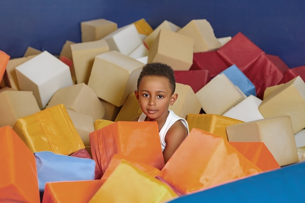 Felice ragazzo afroamericano di otto anni che gioca con i cubi morbidi nella piscina a secco della stanza dei bambini per il compleanno.