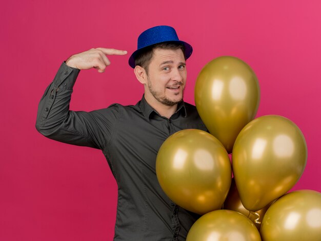 Felice ragazzo giovane partito che indossa il cappello blu che tiene palloncini e punti a se stesso isolato sul rosa