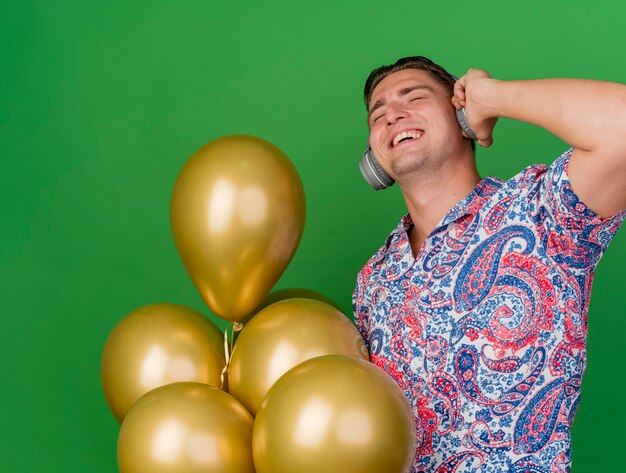 Felice ragazzo giovane partito con gli occhi chiusi che indossa la camicia colorata e cuffie che tengono palloncini isolati su sfondo verde