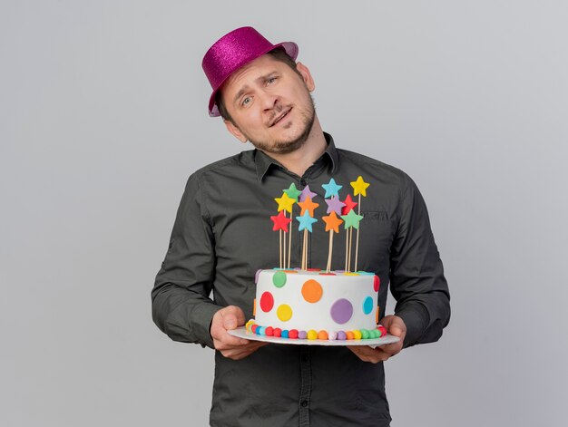 Felice ragazzo giovane partito inclinando la testa indossando il cappello rosa tenendo la torta isolato su sfondo bianco