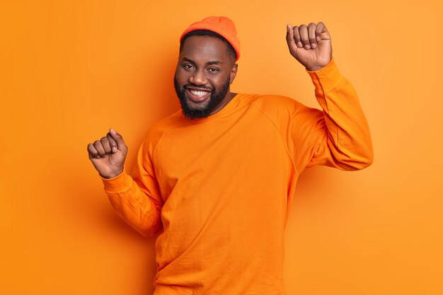 Felice ragazzo spensierato balla sul muro arancione alza le mani si muove attivamente sorride volentieri indossa un cappello alla moda e il ponticello celebra il successo