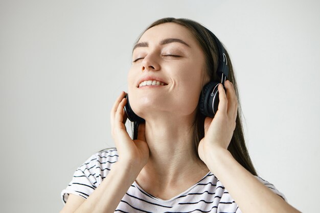 felice rilassata giovane femmina caucasica con i capelli scuri chiudendo gli occhi e gettando la testa indietro, ascoltando musica in cuffia e ballando