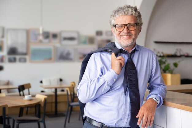 Felice rilassato uomo d'affari maturo in piedi in ufficio caffè, appoggiato al bancone, tenendo la giacca sulla spalla e sorridendo alla telecamera