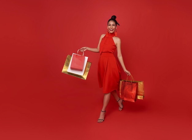 Felice shopaholic asiatico donna che indossa un vestito rosso che tiene il centro commerciale a piedi borsa della spesa