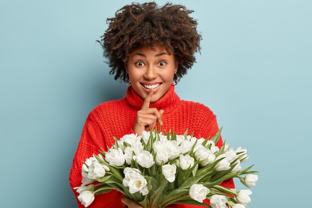 Felice signora afroamericana con i capelli ricci, fa il gesto del silenzio e sorride ampiamente, vestita con un maglione rosso, tiene tulipani bianchi primaverili isolati sopra la parete blu. Non ti dico chi ha presentato i fiori