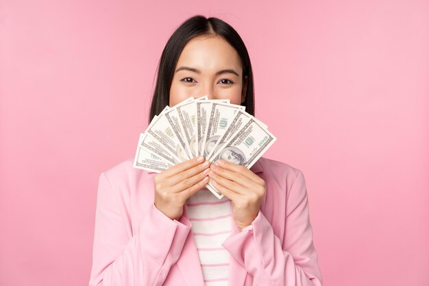 Felice signora asiatica in giacca e cravatta in possesso di dollari di denaro con l'espressione del viso felice in piedi su sfondo rosa