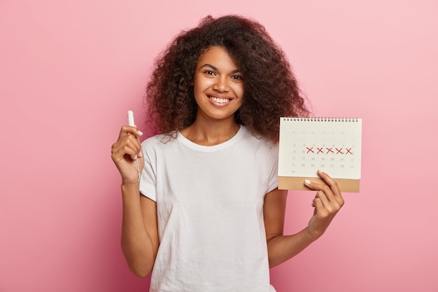 Felice signora dai capelli ricci detiene il calendario delle mestruazioni con contrassegnato giorni di pms e tampone, vestito con una maglietta bianca casual, isolato su sfondo rosa