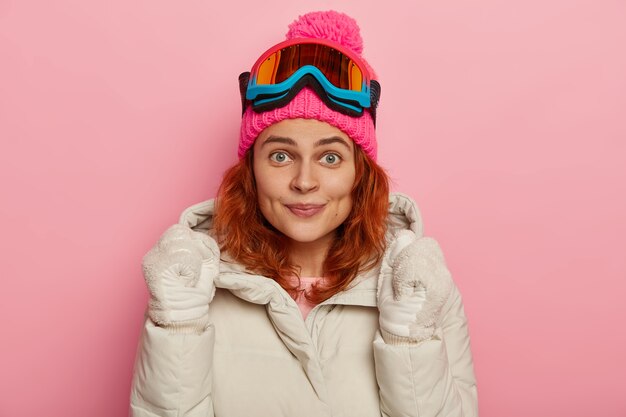 Felice skateboarder alza le mani con espressione esultante, indossa cappotto invernale e guanti, passamontagna, guarda piacevolmente la telecamera, posa contro il muro roseo dello studio.