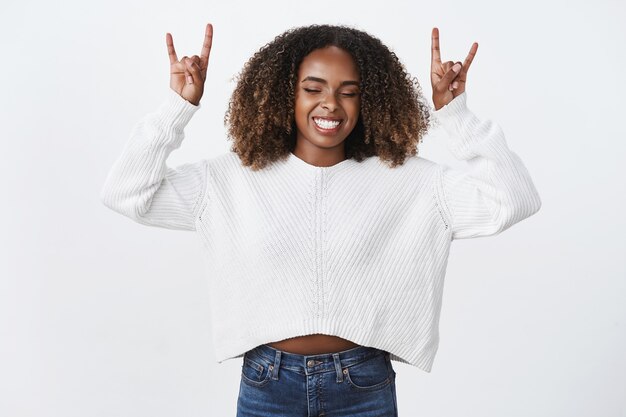 Felice spensierata eccitata donna afroamericana dai capelli ricci chiudere gli occhi entusiasta mostra gesto heavy metal mani in alto divertirsi sorridendo gioiosamente chill intrattenuto, muro bianco