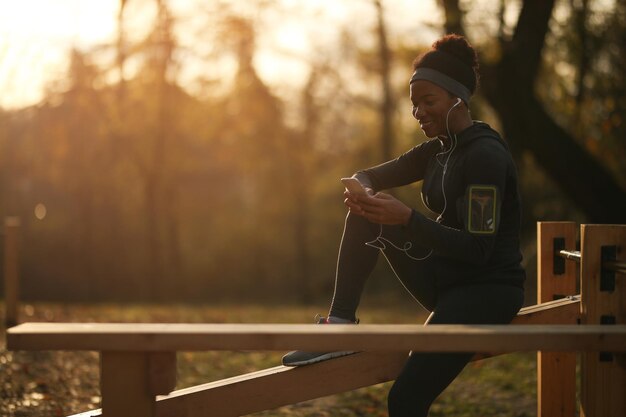 Felice sportiva afroamericana che utilizza lo smartphone mentre si prende una pausa dall'esercizio in natura al tramonto