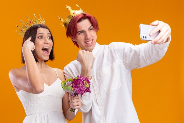 Felice sposi con bouquet di fiori in abito da sposa che indossa corone d'oro sorridendo allegramente facendo selfie utilizzando lo smartphone in piedi sul muro arancione