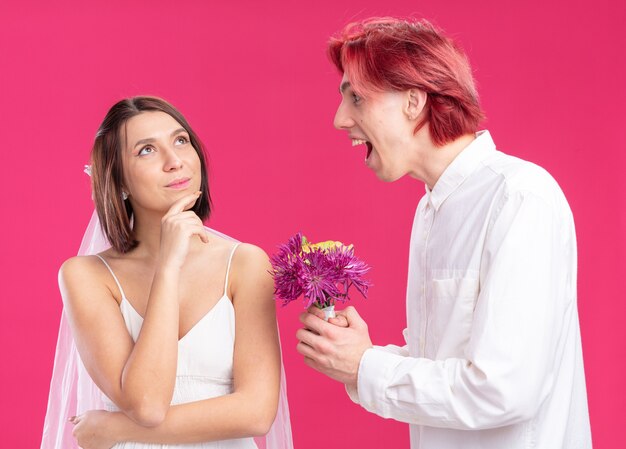 Felice sposi sposo e sposa sposo felice e allegro che dà fiori per la sua sposa sorridente in abito da sposa in piedi sul muro rosa
