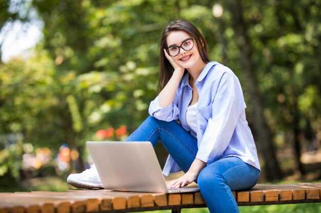 Felice studentessa seduta sulla panchina con il portatile all'aperto nel campus