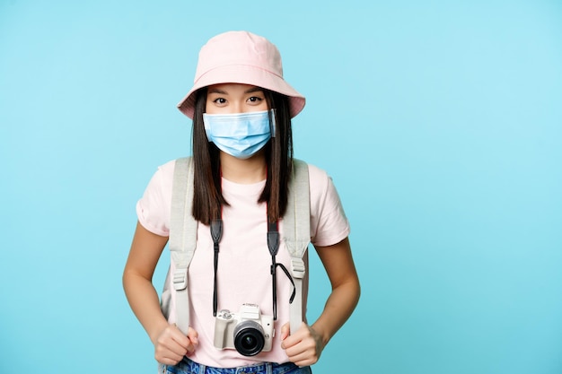 Felice turista asiatica, in piedi in maschera medica, tenendo la macchina fotografica, scattando foto durante le vacanze, visitando la città, esplorando l'estero, in piedi su sfondo blu.