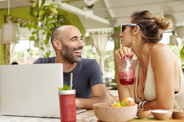 Felice uomo adulto con la barba seduto davanti al laptop aperto, ridendo allegramente, ascoltando la storia della sua ragazza, che sta bevendo un frullato di frutta.