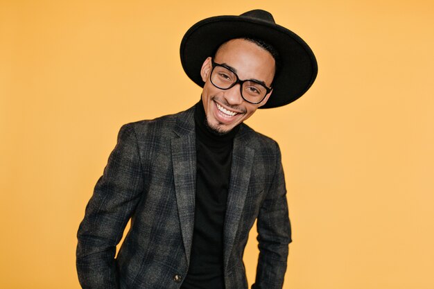 Felice uomo africano in camicia nera e giacca grigia in posa. Foto del ragazzo mulatto positivo con un sorriso sincero isolato sulla parete arancione.