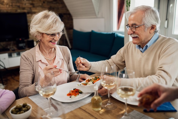 Felice uomo anziano che serve insalata a sua moglie mentre pranza al tavolo da pranzo