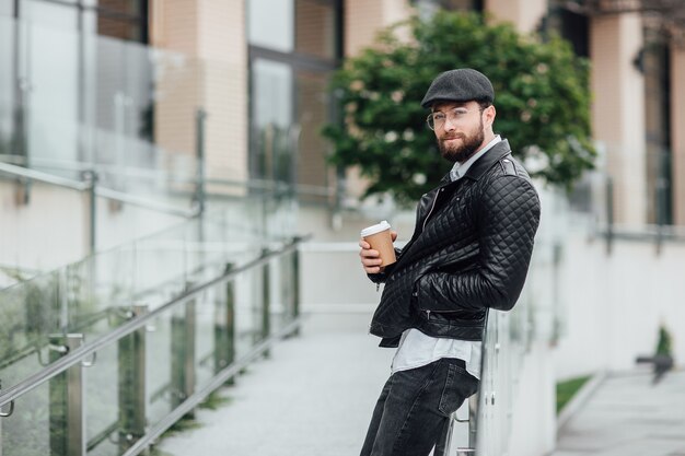 Felice uomo barbuto, serio ed elegante che cammina per le strade della città vicino al moderno centro uffici con caffè