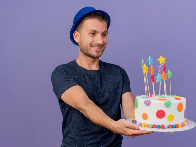 Felice uomo caucasico bello che indossa cappello blu tiene e guarda la torta di compleanno isolata su sfondo viola con spazio di copia