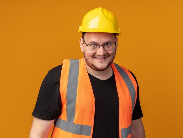 Felice uomo costruttore in giubbotto da costruzione e casco di sicurezza guardando la telecamera sorridendo allegramente in piedi sopra l'arancia