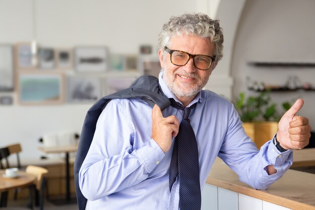 Felice uomo d'affari maturo in piedi in ufficio caffè, appoggiato al bancone, tenendo la giacca sulla spalla, mostrando il pollice in alto o simile