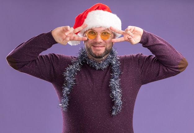 Felice uomo di mezza età che indossa un cappello da Babbo Natale e una ghirlanda di orpelli intorno al collo con gli occhiali che guarda l'obbiettivo che mostra i simboli del segno di v