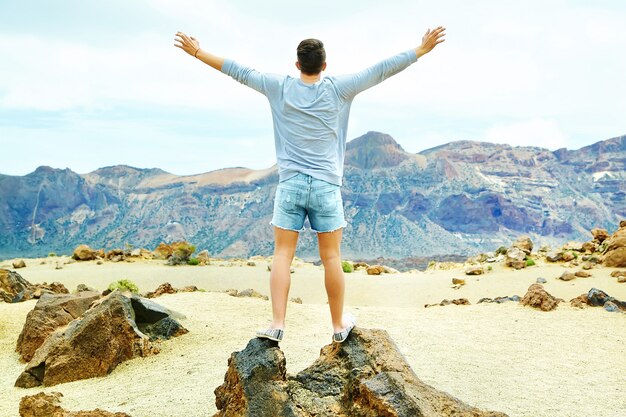 Felice uomo elegante in abiti casual casual in piedi sulla scogliera della montagna con le mani sollevate verso il sole e celebrando il successo