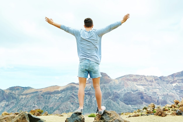 Felice uomo elegante in abiti casual casual in piedi sulla scogliera della montagna con le mani sollevate verso il sole e celebrando il successo