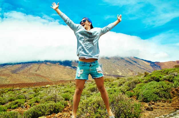 Felice uomo elegante in abiti casual casual saltando davanti alla montagna con le mani sollevate al sole e celebrando il successo