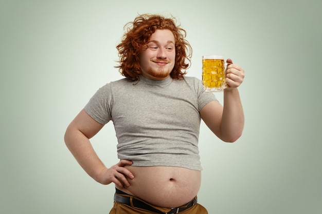 Felice uomo in sovrappeso rossa con la pancia grande che sporge dalla sua maglietta ristretta con in mano un bicchiere di birra fredda, guardando in anticipo, impaziente di sentirne il buon gusto mentre si rilassa a casa dopo il lavoro